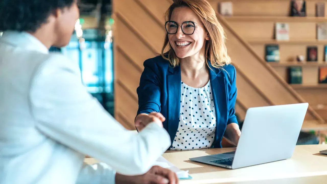 Two professionally dressed women shaking hands while sitting down at desk