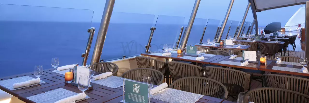 A spacious outdoor dining area on a ship with wooden tables and wicker chairs, each table set with placemats, napkins, wine glasses, and candles. The area is covered by a glass roof and has a clear view of the open ocean in the background.