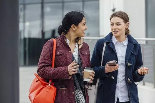 Two women dressed for fall weather walking while holding coffees past an office building