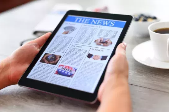 Hands holding a tablet on a coffee table with a the news open