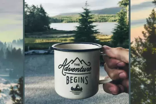 An enamel cup with print is held by a hand against the backdrop of a forest.