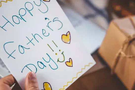 Hand written father's day card being held in front of a table with laptop and wrapped present on it