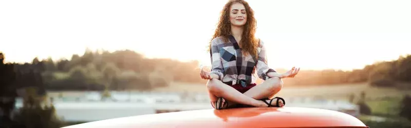 A young woman on a road trip through countryside, sitting on the roof of minivan doing yoga.