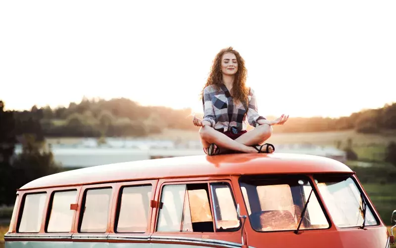 A young woman on a road trip through countryside, sitting on the roof of minivan doing yoga.