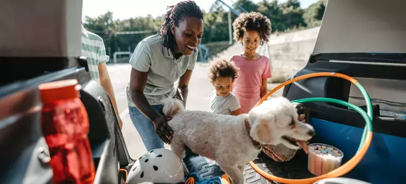 A woman and three children are happily looking at a fluffy white dog in the trunk of a car. The trunk is filled with various items including a hula hoop and sports equipment. It appears to be a bright, sunny day, and they are outdoors.